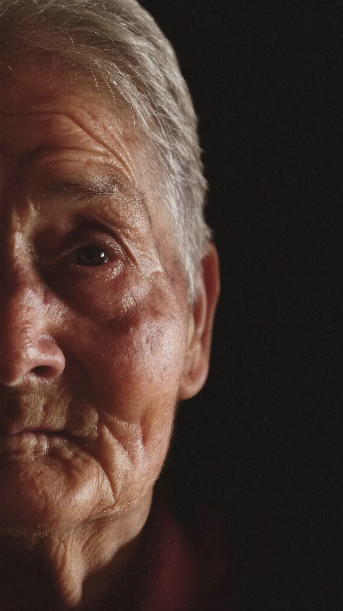 Metade do rosto de uma mulher idosa, iluminado por uma luz baixa, a sair da escuridão.