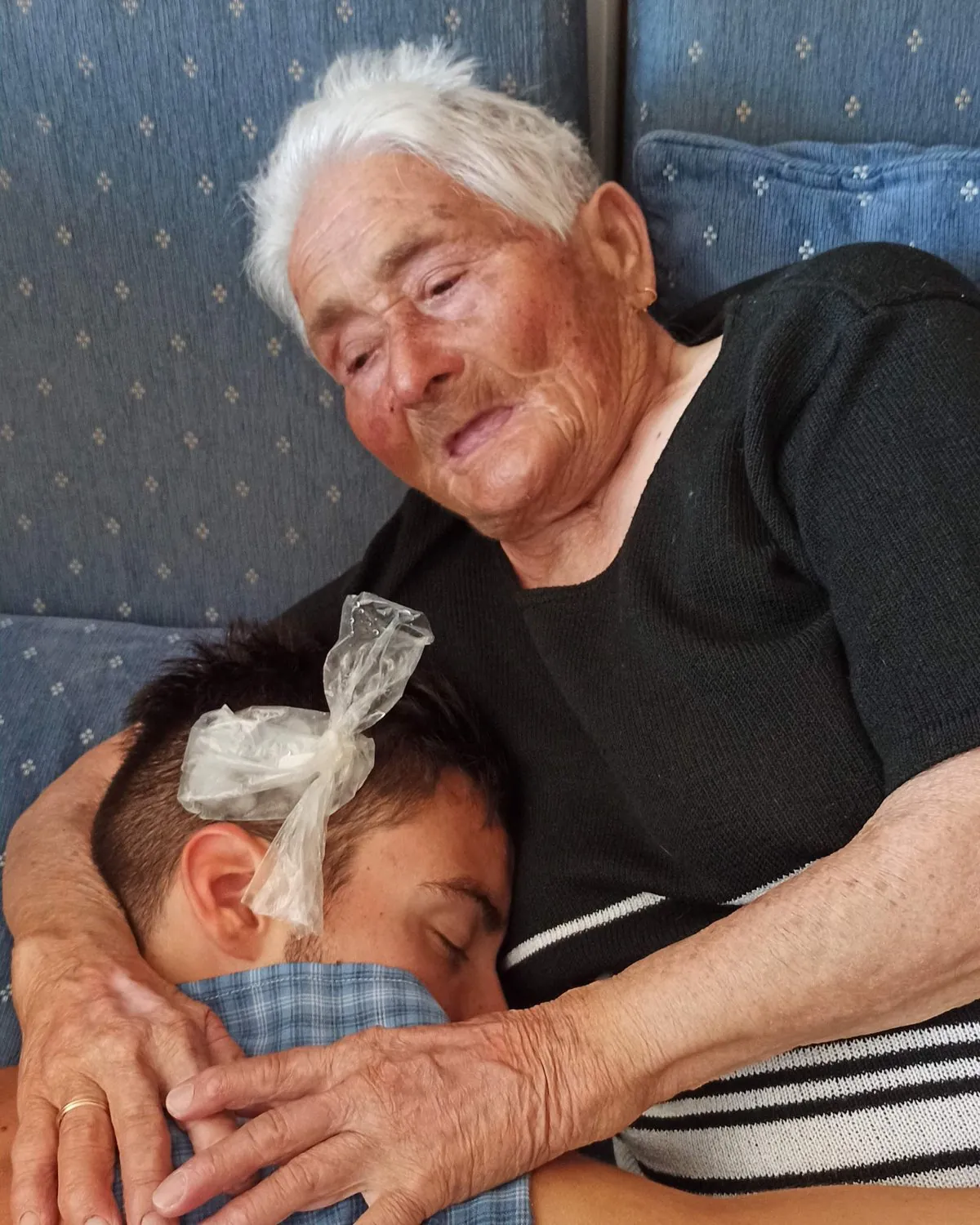 A Maria, de cabelo branco, abraça um neto que dorme encostado a ela, no sofá.