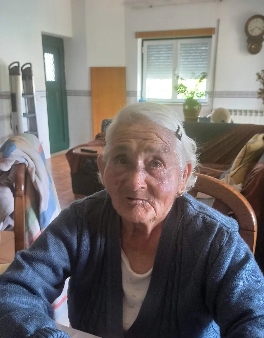 A Maria sentada à mesa da cozinha, de mãos juntas, a olhar para quem a fotografa.